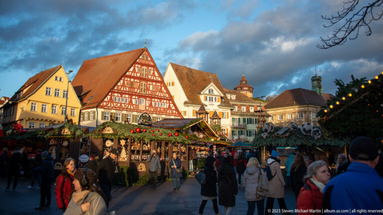 Sunlit gabled and half-timbered houses above a crowded Christmas market decorated with greenery and red ornaments.