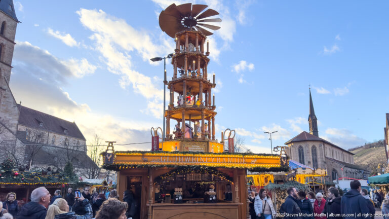 Large tiered wooden Christmas pyramid with rotor above an illuminated market stall and surrounding crowd.