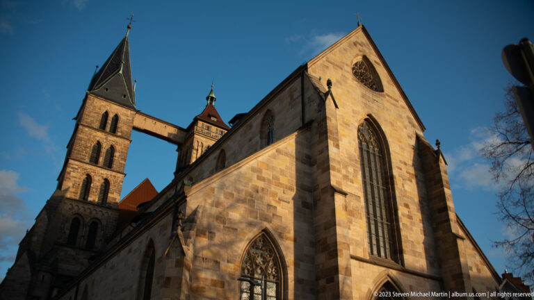 Low oblique view of Stadtkirche St. Dionys with tall pointed window, square tower, and elevated bridge between towers.