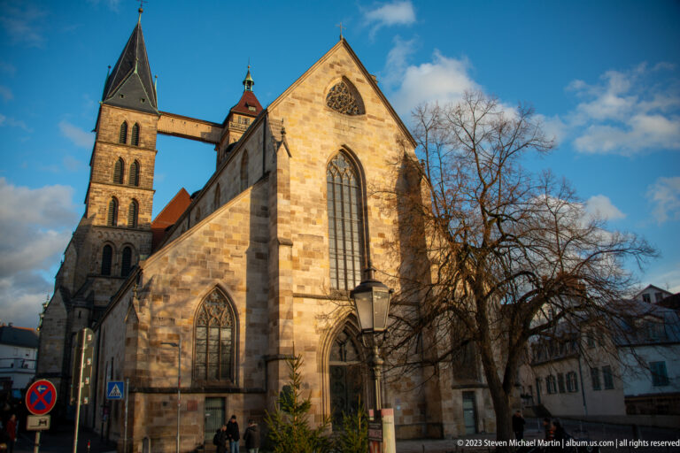Stone gabled end of Stadtkirche St. Dionys with tall Gothic window, square tower, connecting bridge, and leafless tree.