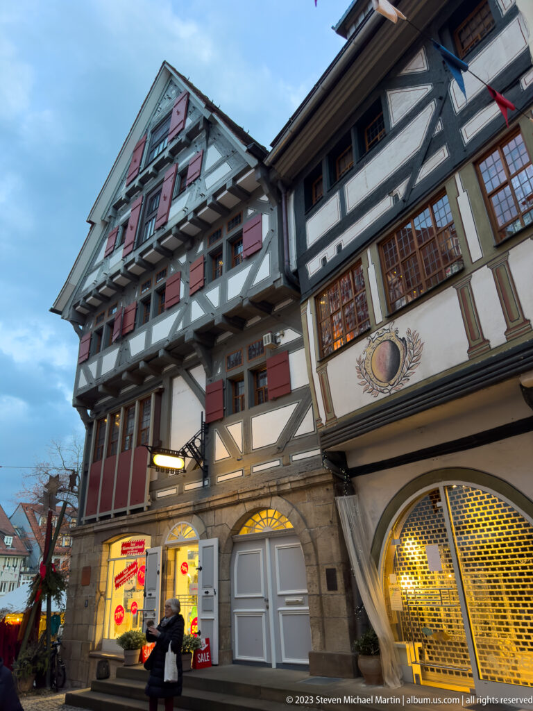 Low-angle view of a multi-story half-timbered building with red shutters, projecting bays, and modern shops at street level.