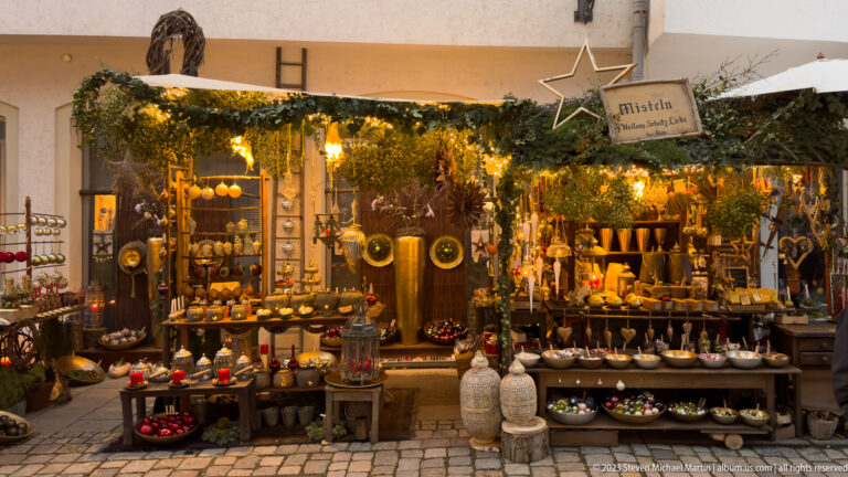 Warmly lit Christmas market stall displaying pottery, bowls, lanterns, small sculptures, and hanging decorations beneath evergreen garlands.