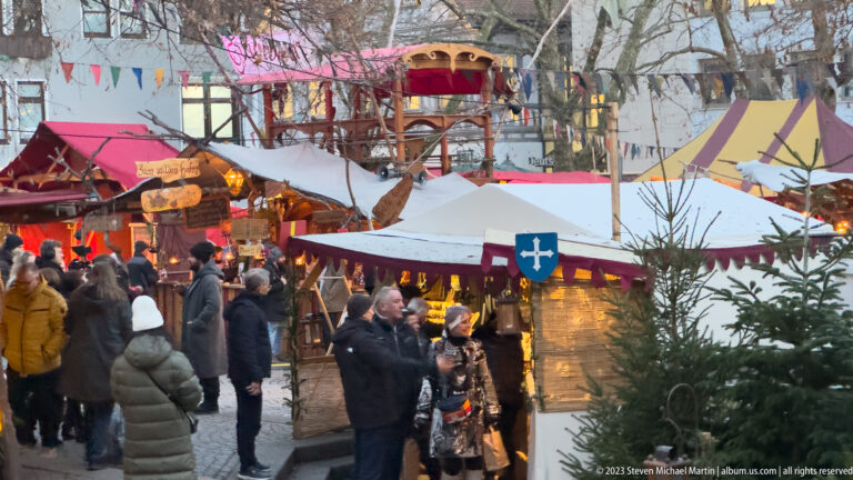 Crowded medieval-themed market with canvas tents, timber booths, heraldic shields, colorful awnings, and bare trees.