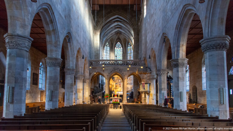 Central nave of Stadtkirche St. Dionys with wooden pews, pointed arcades, stone piers, gallery, and illuminated chancel.