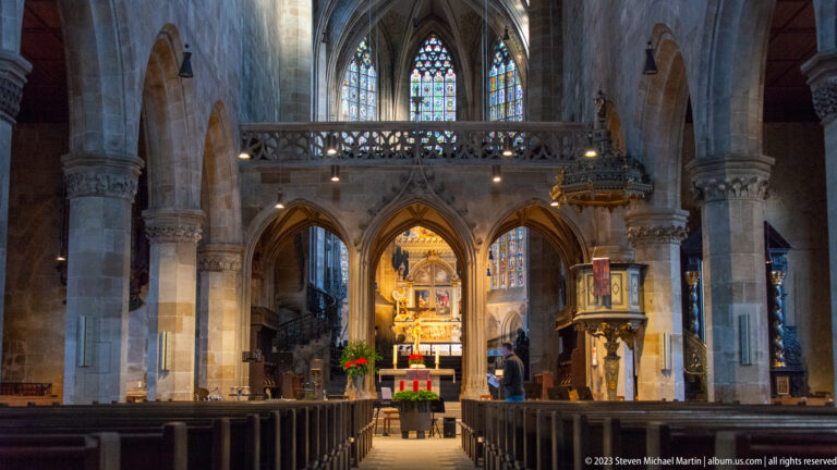 Chancel of Stadtkirche St. Dionys with pointed arches, traceried gallery, illuminated altar, carved pulpit, and stained glass.
