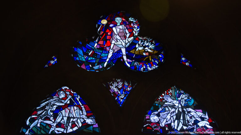 Modern figurative stained glass in vivid blue, red, white, and green glowing within dark Gothic tracery.