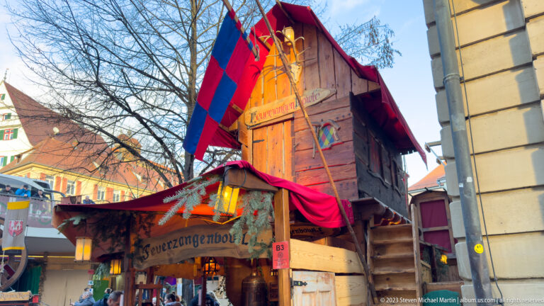 Tall rustic wooden Feuerzangenbowle stall draped in red and blue fabric with an exterior stair.