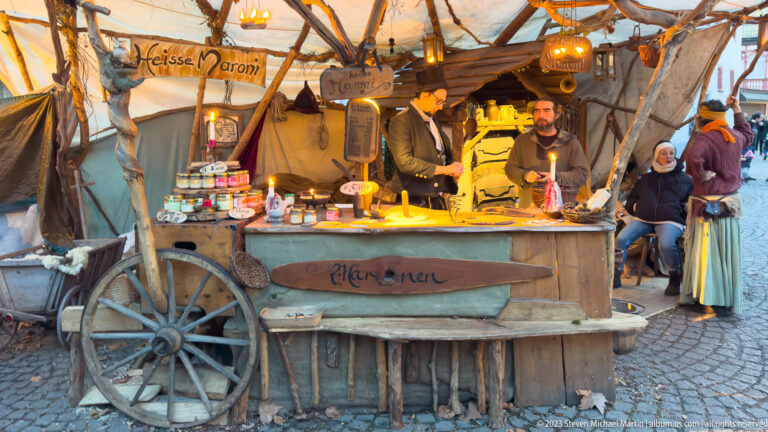 Rustic Alchemie market booth with bearded vendor, jars, bottles, candles, branches, canvas, and wagon wheels.