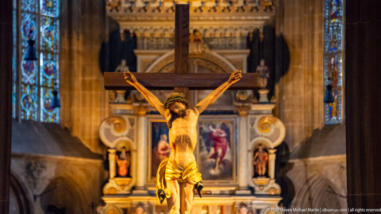 Large crucifix in sharp focus before an ornate painted altar and stained-glass windows inside a historic church.
