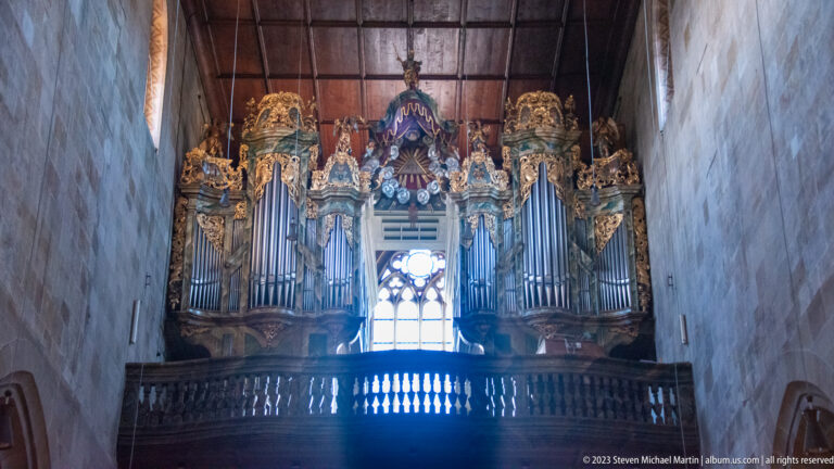 Large ornate pipe organ with silver pipes, gilded carving, and sculptural decoration on a church gallery beneath a timber ceiling.