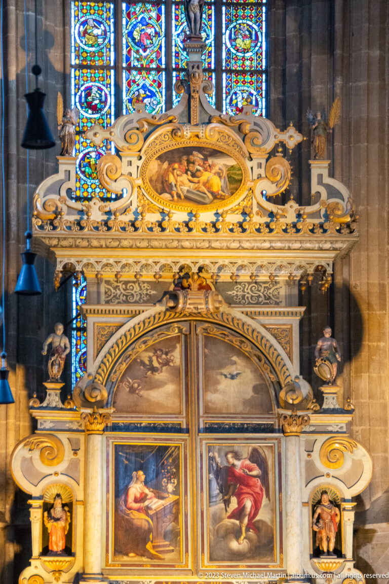 Ornate church altarpiece with painted religious panels, gilded scrollwork, small sculpted figures, and stained glass behind.