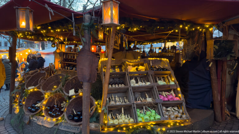 Warmly lit rustic market booth with shelves of handmade goods, wreaths, branches, baskets, and canvas canopy.