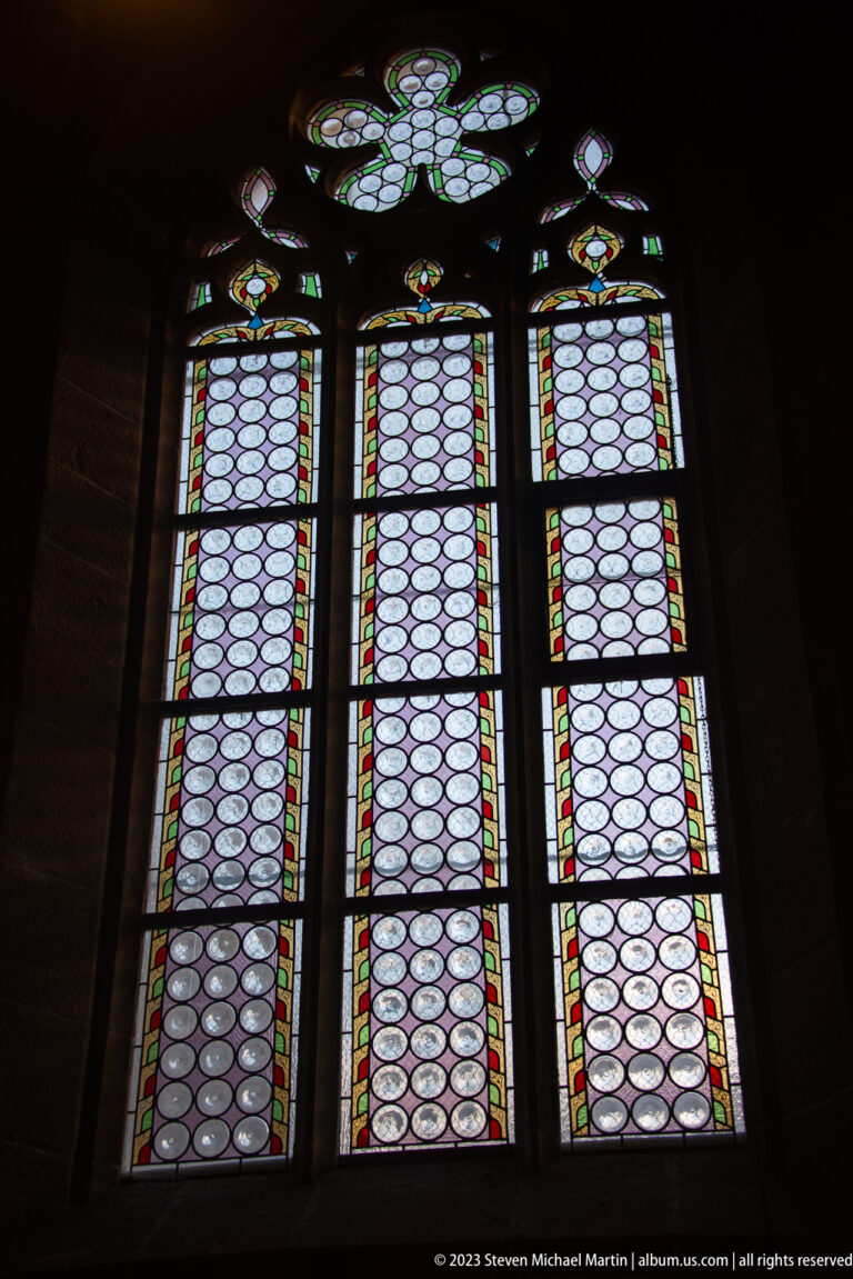 Three-light Gothic window with pale circular glass quarries, colored borders, and decorative tracery glowing in a dark interior.