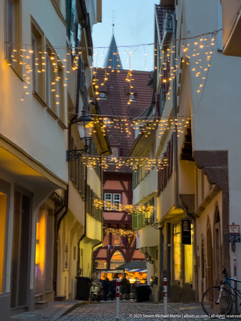 Narrow old-town street with overhead Christmas lights, shuttered façades, tiled roofs, and half-timbered buildings in the distance.