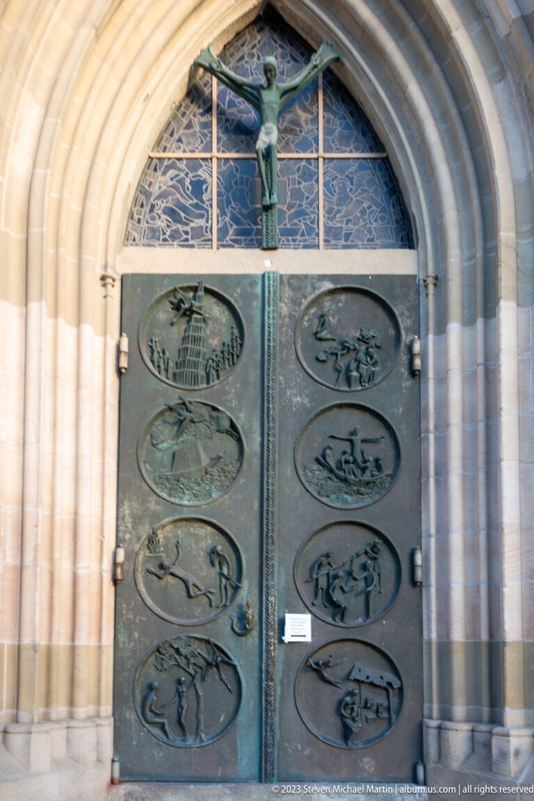 Bronze double doors with oval relief panels beneath a pointed Gothic stone arch and small crucifix.