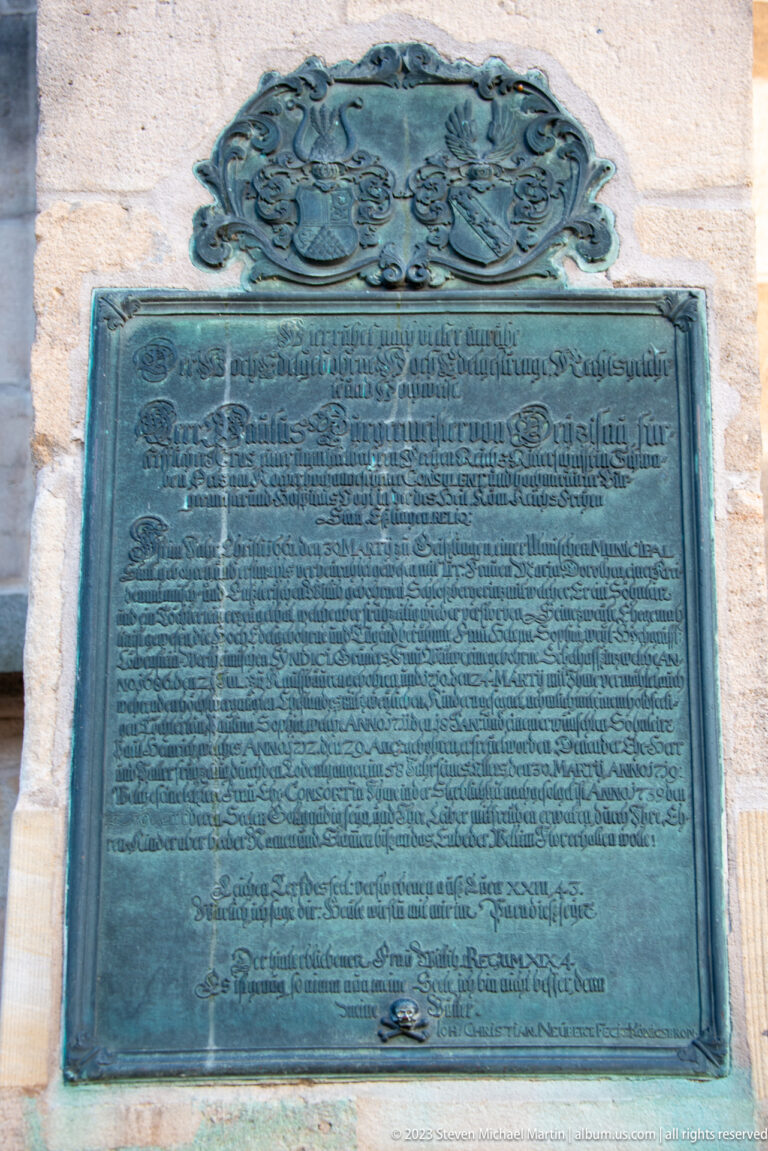 Large patinated bronze plaque with dense German inscription and ornate crest-like decoration mounted on pale stone.