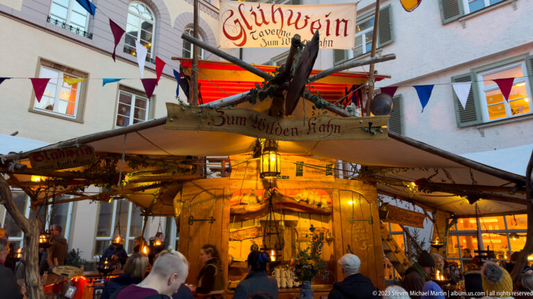 Busy timber market stall labeled Zum Wilden Hahn, decorated with branches, bird figure, pennants, red awnings, and warm lights.