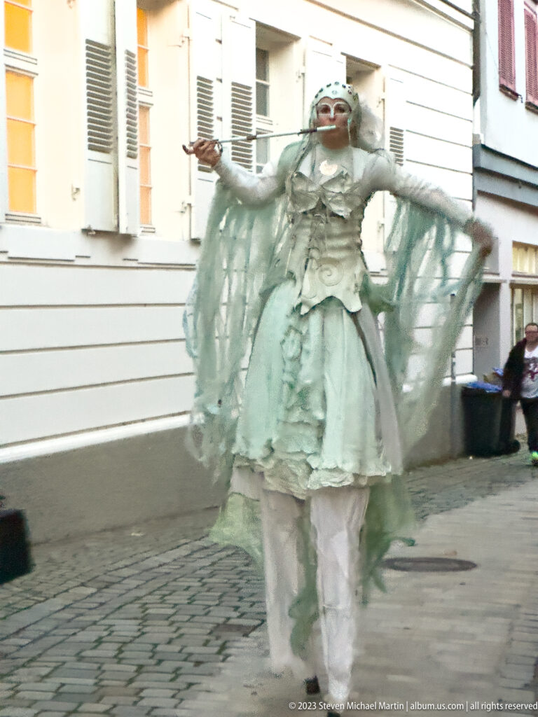 White-costumed street performer with extended arms, pale face, layered skirt, and flowing translucent fabric on a cobbled street.