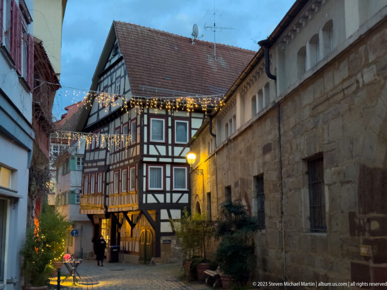 Narrow cobbled lane with projecting half-timbered house, long stone wall, warm lamps, and small Christmas tree.