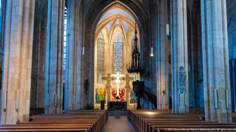 Central aisle through a Gothic church nave with wooden pews, tall stone piers, carved pulpit, stained glass, and illuminated chancel.