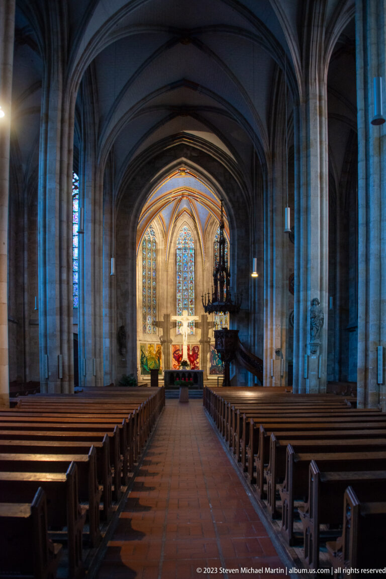 Wide axial view of a Gothic nave with ribbed vaults, wooden pews, tall piers, and brightly illuminated chancel.