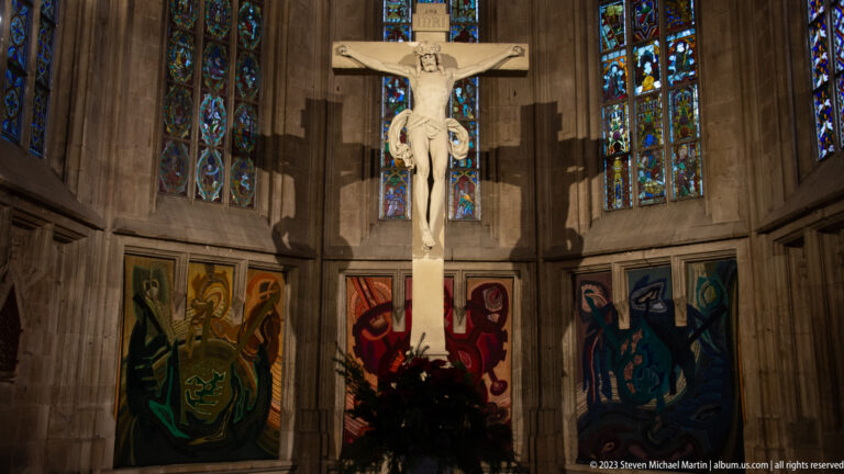 Large pale crucifix centered before tall stained-glass windows and colorful textile panels in a Gothic chancel.