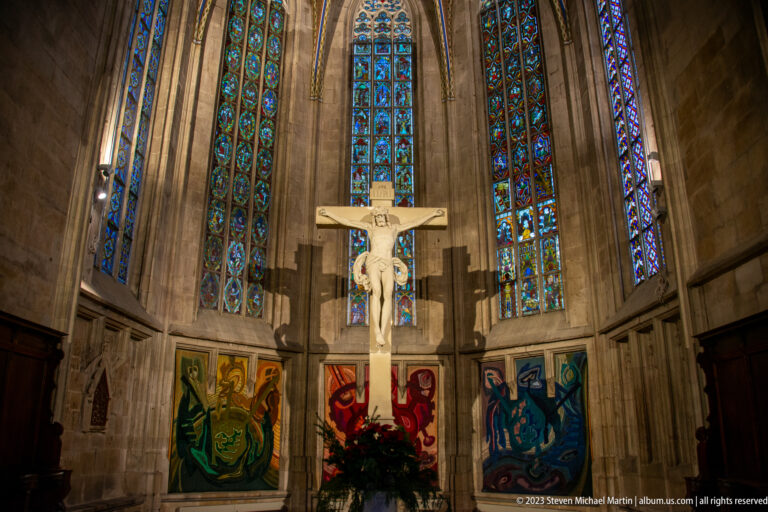 Crucifix beneath a fan of tall stained-glass lancets with colorful textile panels and flowers around the Gothic chancel.