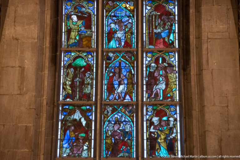 Three Gothic lancets filled with nine colorful figurative stained-glass scenes arranged in stacked narrative panels.