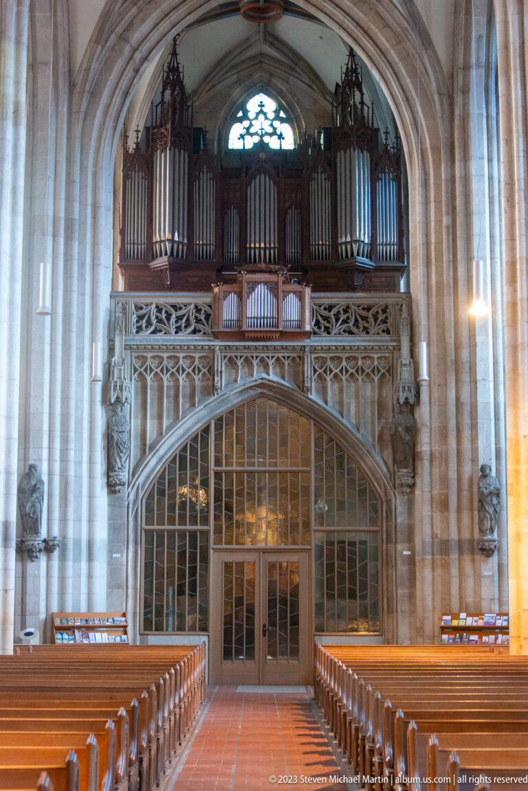 Large pipe organ above an ornate stone gallery and glazed screen at the west end of a Gothic church.