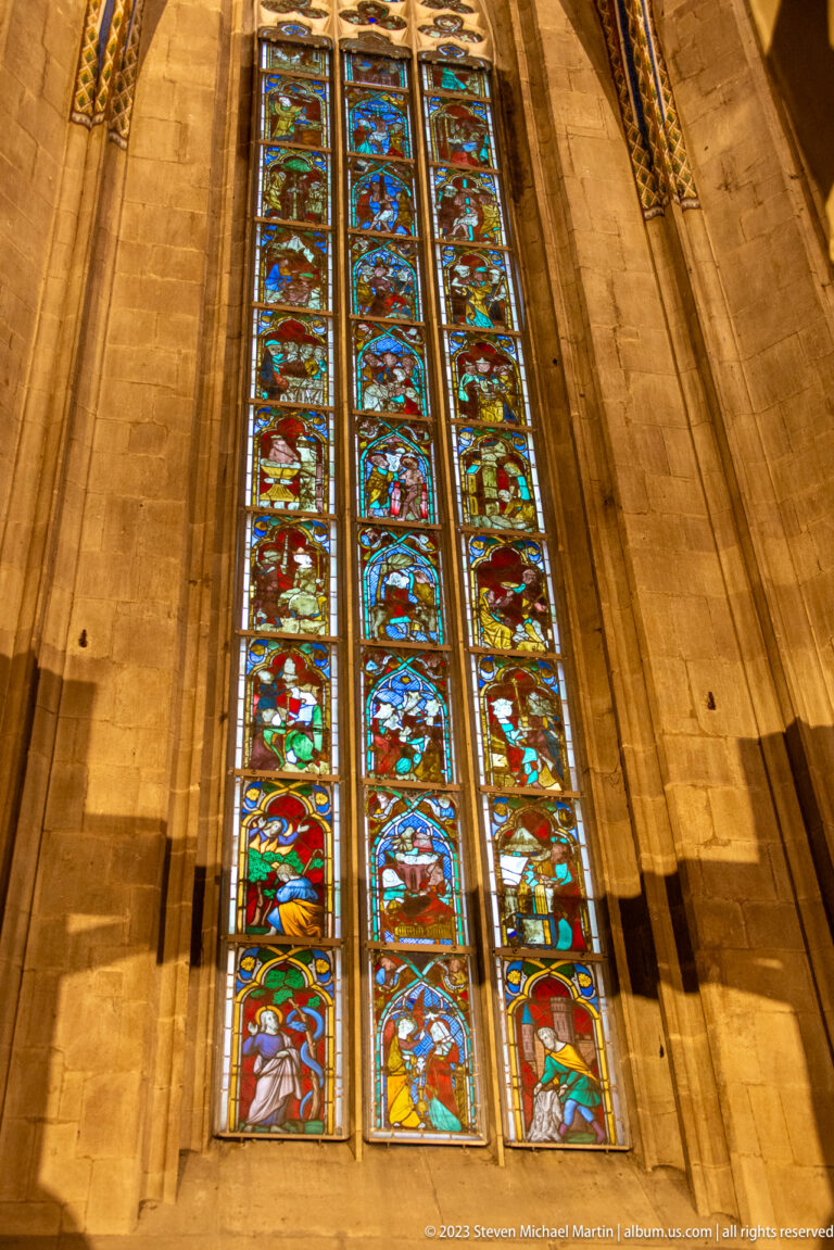 Upward view of tall three-light stained-glass window with stacked colorful figurative scenes set in pale stone.