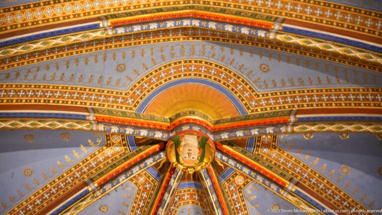 Close angled view of a richly painted Gothic vault with multicolored ribs, gold stars, patterned borders, and small central medallion.