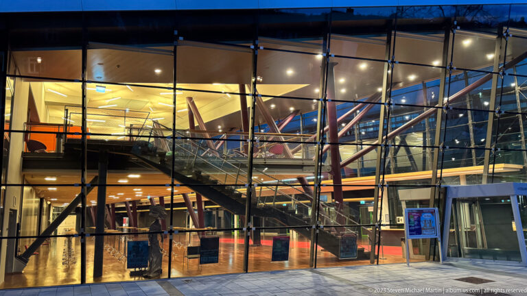 Glass-fronted modern public building revealing illuminated interior floors, angular staircases, and diagonal structural members at dusk.