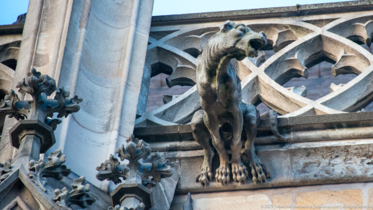 Slender long-necked stone grotesque standing upright before looping Gothic tracery and clustered crockets.