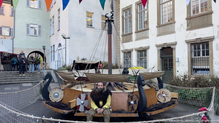 Bearded interpreter seated before a small wooden boat with mast, rigging, oars and painted round shields.