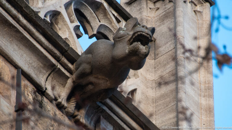 Open-mouthed stone gargoyle projecting from a Gothic church corner beneath deep openwork tracery.