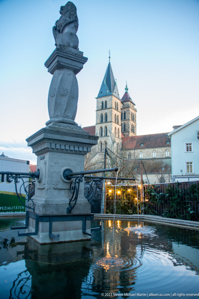 Stone fountain basin and sculpted pier in the foreground with the twin-towered Stadtkirche St. Dionys beyond.