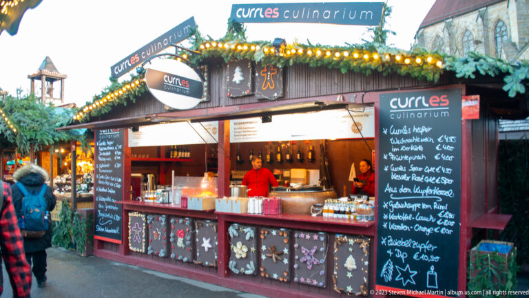 Curles Culinarium Christmas market booth with blackboard menus, bottles, warm lights, greenery, and a vendor in red.