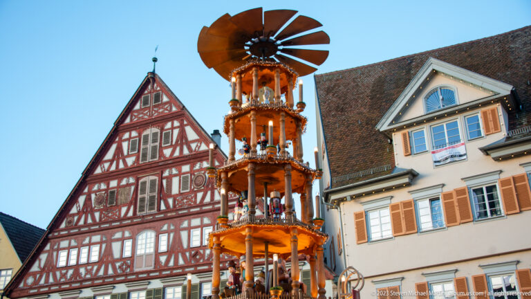 Illuminated wooden Christmas pyramid standing before a red-and-white half-timbered gabled house and pale shuttered building.