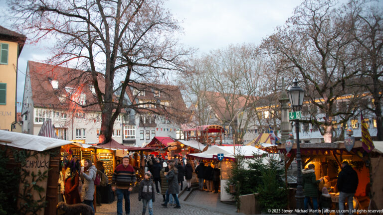 Busy Christmas market with visitors, canvas-roofed stalls, warm lights, bare trees, and historic houses at dusk.