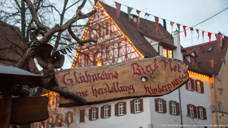 Hand-painted wooden sign listing Glühweine, beer, mead, hot juice, and children’s punch before a half-timbered house.