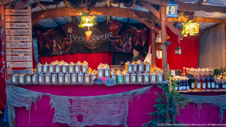 Beerenweine market stall with rows of ceramic and glass bottles, red drapery, rustic lanterns, and hand-lettered signs.