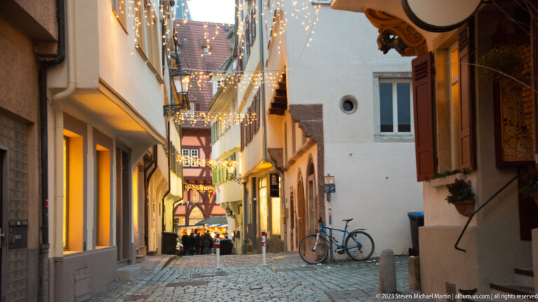 Narrow cobbled old-town alley with overhead Christmas lights, shuttered façades, bicycle, and brighter market activity ahead.