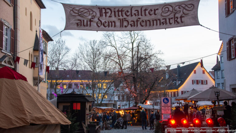 Banner reading Mittelalter auf dem Hafenmarkt above a crowded market street with tents, stalls, pennants, and bare trees.