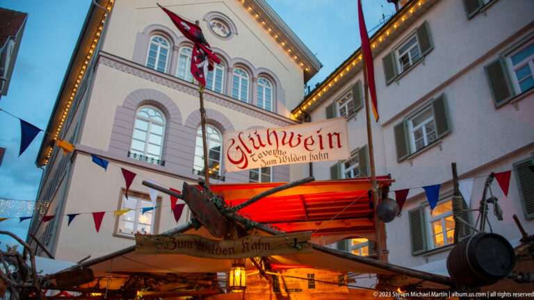 Glowing Zum Wilden Hahn Glühwein booth with red canopy and pennants beneath a pale historic building with arched windows.