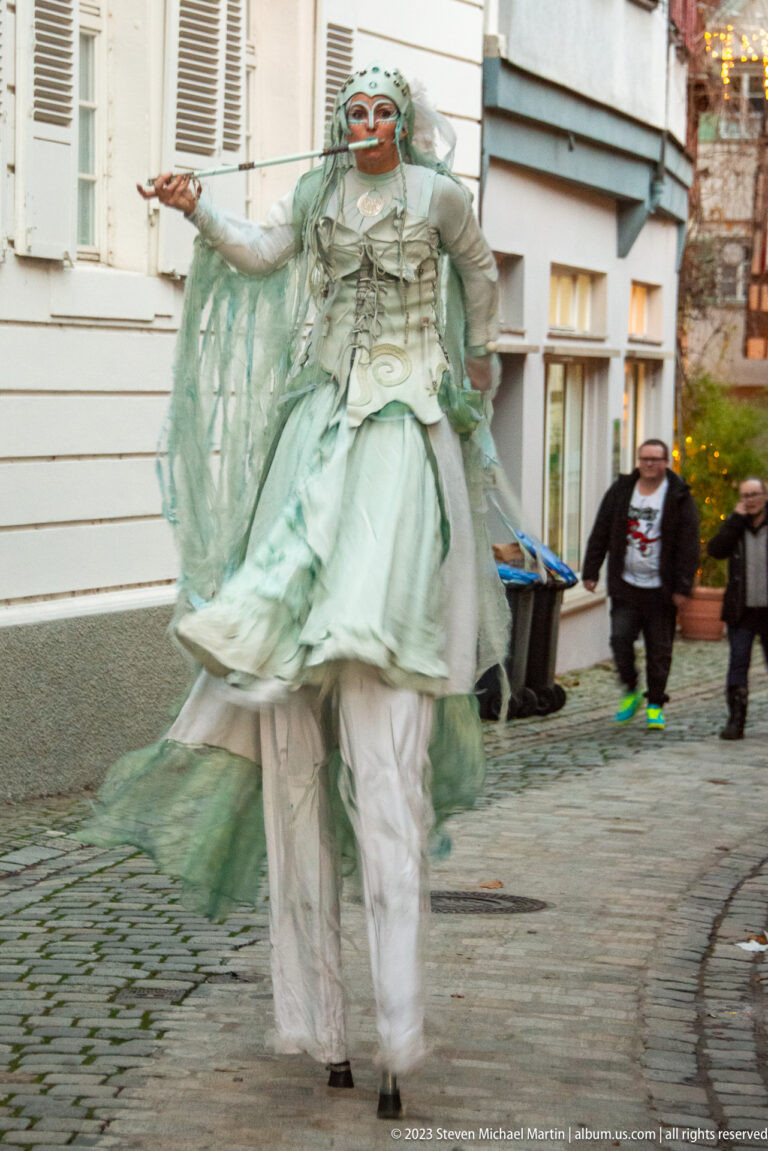 Performer in layered white and pale-green costume moving across cobblestones, with flowing fabric and a stylized pale face.