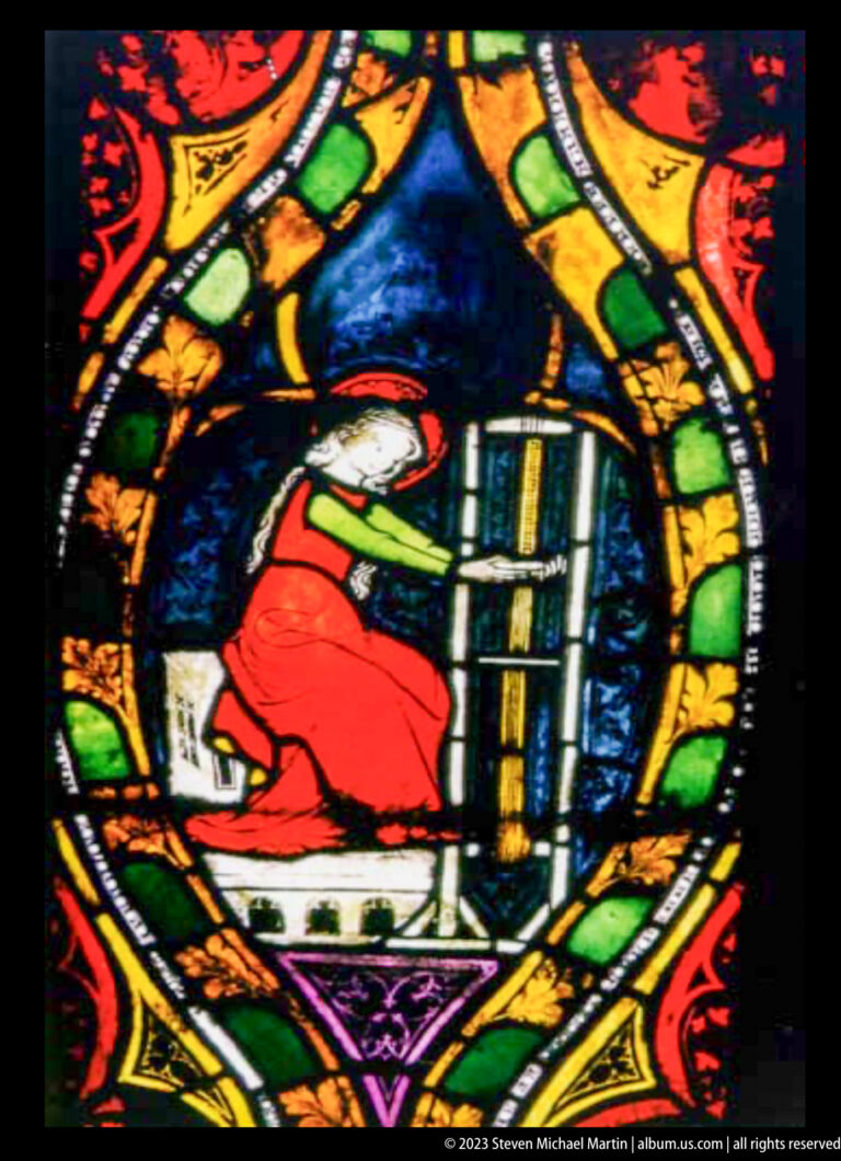 Medieval stained-glass roundel showing a robed figure reading from an open book on a tall lectern.