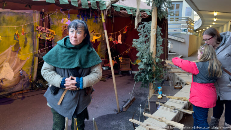 Woman in green cape and medieval-style clothing speaking with visitors at a small craft stall.