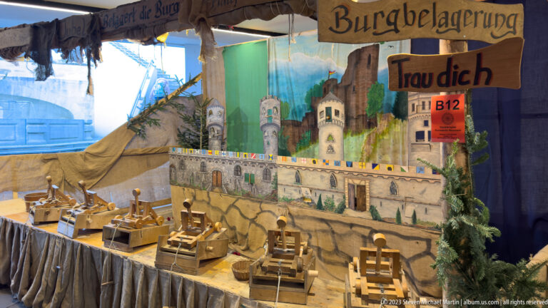 Tabletop castle-siege game with painted fortress, small wooden launchers and a Burgbelagerung sign.