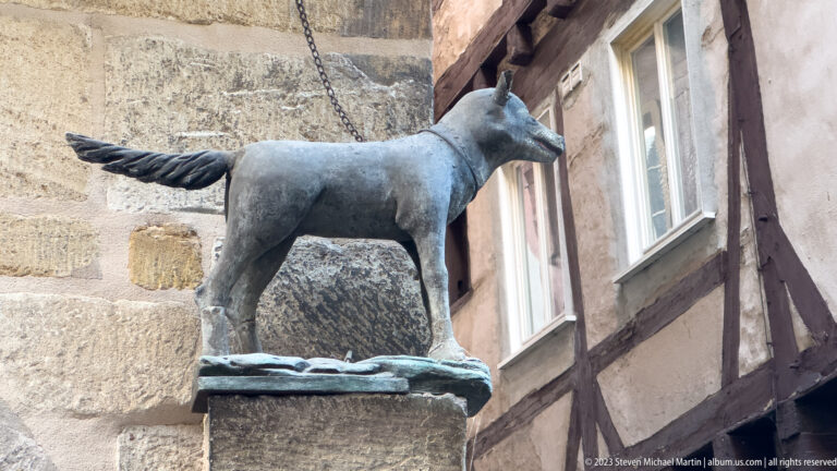 Small metal horse- or donkey-like sculpture on a stone pedestal beside a half-timbered wall.