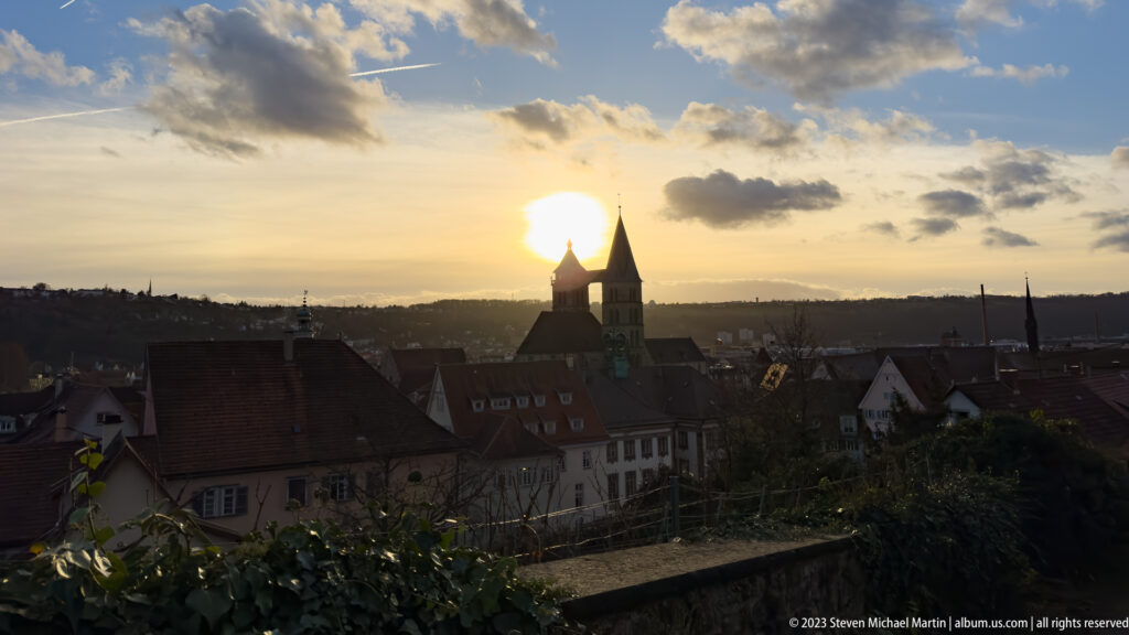 Sunset over Esslingen rooftops with the twin towers of Stadtkirche St. Dionys silhouetted near the sun.